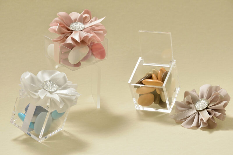 Cubo portaconfetti in plexiglass cm. 5 x 5 x 5 e scatolina in PVC alimentare cm. 6 x 6 x 3, abbelliti da una margherita dai petali di raso, con pistillo glitterato. I confetti sfumati lilla, turchese e marrone, ripropongono rispettivamente, il tema della primavera, del mare e dell’autunno.