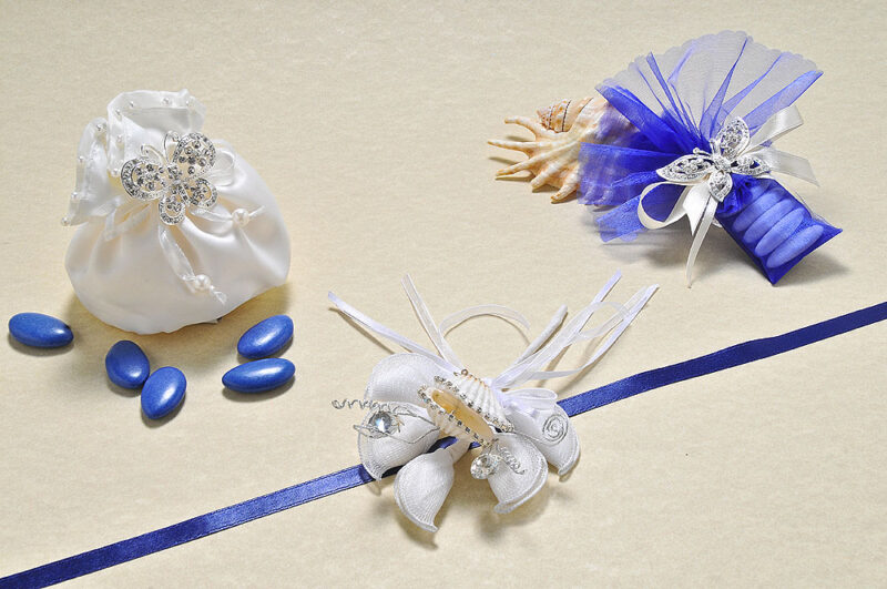 Sacchetto bombato in raso avorio con perle e tubino velato blu, impreziositi da ciondolo con farfalla argentata con strass. Un tocco luminoso il bouquet con la conchiglia vera contornata da strass.